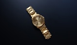 A gold wristwatch with a metallic band is centered against a dark background. The watch has a round face with minimalistic hour markers and hands.