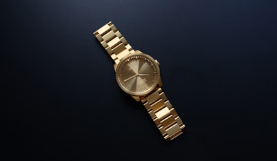 A gold wristwatch with a metallic band is centered against a dark background. The watch has a round face with minimalistic hour markers and hands.