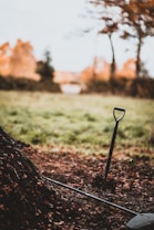 A garden shovel is stuck in a pile of soil or leaves with a rake resting nearby. The area is surrounded by a grassy field and trees, suggesting an outdoor setting in autumn.