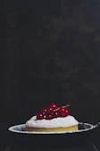 A small, round dessert dusted with powdered sugar is topped with bright red berries and placed in a dark, shallow pan. The background is dark and slightly textured, putting full focus on the dessert.