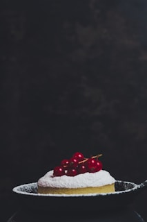 A small, round dessert dusted with powdered sugar is topped with bright red berries and placed in a dark, shallow pan. The background is dark and slightly textured, putting full focus on the dessert.