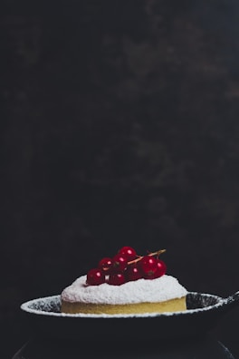 A small, round dessert dusted with powdered sugar is topped with bright red berries and placed in a dark, shallow pan. The background is dark and slightly textured, putting full focus on the dessert.