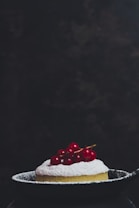 A small, round dessert dusted with powdered sugar is topped with bright red berries and placed in a dark, shallow pan. The background is dark and slightly textured, putting full focus on the dessert.