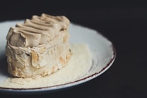 A piece of creamy dessert, possibly tiramisu or a mocha-flavored cake, is plated elegantly. The dessert is garnished with thinly sliced almonds or similar nut flakes. It sits on a round, white plate with a rustic, worn rim, creating a contrast with the dark background.