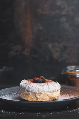 A pastry garnished with almond slices is placed on a dark plate. The top of the pastry is lightly dusted with powdered sugar and cocoa powder, and it is adorned with a couple of dark berries. In the background, a glass jar with a metallic lid is partially visible.
