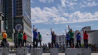 A group of people stand on raised platforms in an urban setting with their fists raised. They are wearing brightly colored shirts of various hues, and the scene is set against the backdrop of modern buildings and a partly cloudy sky. Numerous bicycles and additional people are visible in the background.