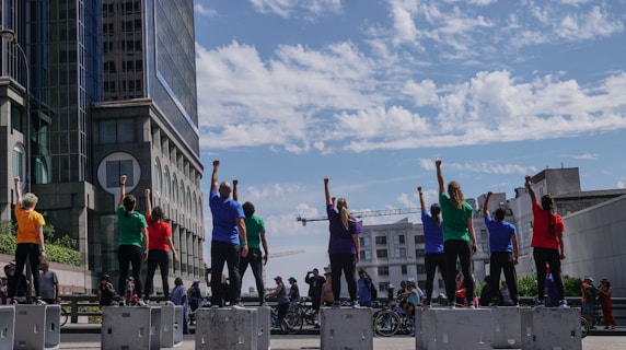 A group of people stand on raised platforms in an urban setting with their fists raised. They are wearing brightly colored shirts of various hues, and the scene is set against the backdrop of modern buildings and a partly cloudy sky. Numerous bicycles and additional people are visible in the background.