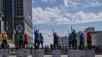 A group of people stand on raised platforms in an urban setting with their fists raised. They are wearing brightly colored shirts of various hues, and the scene is set against the backdrop of modern buildings and a partly cloudy sky. Numerous bicycles and additional people are visible in the background.