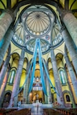 A grand and ornate church interior with tall stone columns and arches, adorned with long blue drapes hanging from the dome. Stained glass windows and intricate architectural details are visible. Chairs are aligned in rows facing the altar, where a person stands in contemplation.