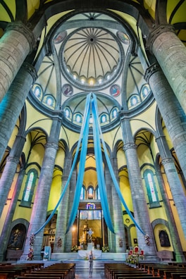 A grand and ornate church interior with tall stone columns and arches, adorned with long blue drapes hanging from the dome. Stained glass windows and intricate architectural details are visible. Chairs are aligned in rows facing the altar, where a person stands in contemplation.