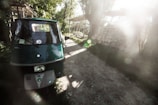 A green three-wheeled vehicle is parked on a dirt path surrounded by lush greenery. Sunlight filters through the trees, casting soft, dreamy light across the scene. In the background, there is a stack of neatly piled logs.