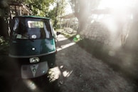 A green three-wheeled vehicle is parked on a dirt path surrounded by lush greenery. Sunlight filters through the trees, casting soft, dreamy light across the scene. In the background, there is a stack of neatly piled logs.