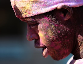 A person with dark skin is shown in profile, covered in vibrant colored powders typically associated with a festival, such as Holi. The face and clothing are heavily smeared with pink, yellow, and other hues, creating a festive and lively appearance. The person is wearing a headband that is also dusted with colored powder.