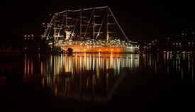 A large, illuminated cruise ship is docked at night, with strings of lights outlining its masts. The lights create a warm glow, reflecting on the calm, dark water, adding to the serene atmosphere. Surrounding the ship are gentle silhouettes of the shoreline and sparse, distant lights.