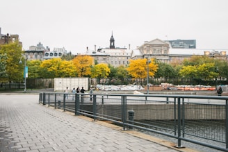 A paved walkway by a riverside with a metal railing in the foreground. In the background, there is a historic, multi-story stone building surrounded by trees with autumn foliage. People are gathered near a white storage container, and various materials are covered in tarps on the ground.