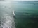 A sailboat with a white sail glides smoothly over the shimmering green water. In the distance, a small motorboat and kayakers can be seen moving across the sea under a clear sky. Sunlight reflects vividly on the surface, creating a sparkling effect.
