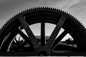 A large, industrial gear with visible teeth dominates the composition against a partially cloudy sky. The gear appears to be part of a heavy machinery or equipment, with other mechanical components visible in the background. The image is in black and white, giving it a stark, dramatic feel.
