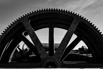 A large, industrial gear with visible teeth dominates the composition against a partially cloudy sky. The gear appears to be part of a heavy machinery or equipment, with other mechanical components visible in the background. The image is in black and white, giving it a stark, dramatic feel.