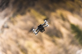 A small drone with four rotors is captured mid-flight against a blurred, natural background. The drone's camera is prominently visible, and the motion of the propellers suggests it's actively flying.