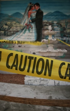 A mural depicting a bride and groom in a wedding scene with a landscape background is visible. In front of the mural, there is yellow caution tape with the word 'CAUTION' written on it.