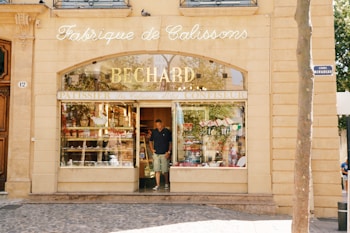 An elegant storefront with 'Fabrique de Calissons Bechard' inscribed above the entrance. The shop has large windows displaying various confectioneries. A person is seen exiting the store. The architecture features stone walls and decorative elements.