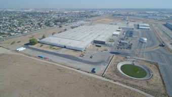 Aerial view of a large industrial warehouse complex surrounded by dry, open land and adjacent urban area. The facility includes multiple parking lots and loading docks. A water tower and a small circular body of water are visible nearby.