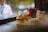 On a countertop, several burgers with fresh lettuce and buns rest, surrounded by a slightly blurred background. Condiment bottles and paper cups can be seen in the background, creating a busy and casual dining atmosphere.