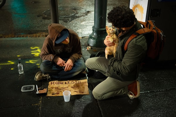 Man kneels next to a homeless man, sharing a meal and conversation with him.