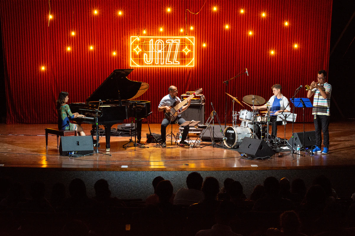赤いカーテンと黄色いネオンの「JAZZ」の文字を背にした舞台で、左から、グランドピアノを弾く長い髪の女性、ベースを弾く男性、ドラムを叩く男性、トランペットを吹く縞シャツの男性の4人が演奏している。手前の客席には観客の後ろ姿のシルエットが並ぶ