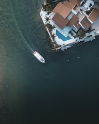 An aerial view of a luxury waterfront property with a contemporary architectural style featuring geometric shapes and a terracotta-colored roof. A sleek white boat navigates the adjacent dark green water. A rectangular swimming pool is visible in the backyard, surrounded by palm trees and a patio.