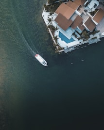 An aerial view of a luxury waterfront property with a contemporary architectural style featuring geometric shapes and a terracotta-colored roof. A sleek white boat navigates the adjacent dark green water. A rectangular swimming pool is visible in the backyard, surrounded by palm trees and a patio.