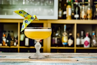 A cocktail in an elegant glass, topped with a colorful origami bird, is placed on a wooden surface. Behind the drink, shelves stocked with various liquor bottles create a blurred background.