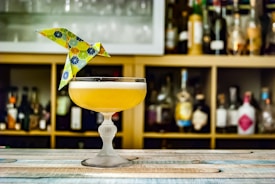 A cocktail in an elegant glass, topped with a colorful origami bird, is placed on a wooden surface. Behind the drink, shelves stocked with various liquor bottles create a blurred background.
