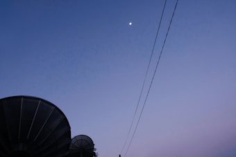A twilight sky with a small moon is visible above two large satellite dishes. Power lines stretch diagonally across the scene, adding a sense of depth.