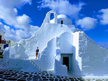 A stunning whitewashed building with traditional Cycladic architecture, featuring domes and smooth, curved surfaces. The building is set against a vibrant blue sky filled with fluffy white clouds. A person stands on the stone-paved steps of the building, adding scale and life to the scene.