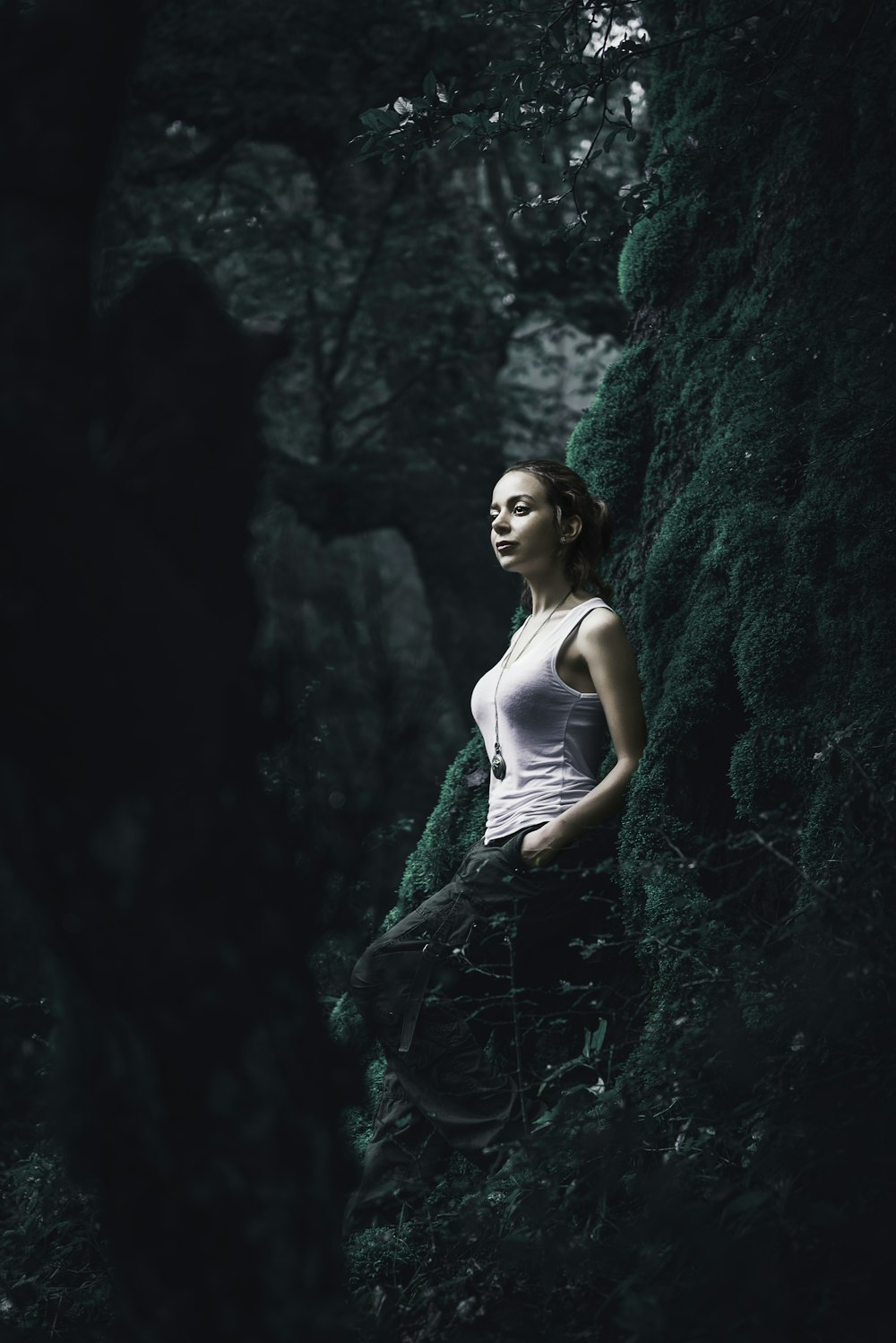 woman in white tank top stands while leaning on green moss covered tree trunk