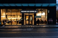A coffee shop with large glass windows showing an inviting interior. Several people are seated at tables, enjoying their drinks, while others can be seen standing inside. The establishment is lit with warm lighting that highlights artwork on the walls and the interior decor.