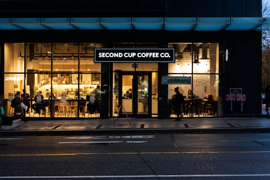 A coffee shop with large glass windows showing an inviting interior. Several people are seated at tables, enjoying their drinks, while others can be seen standing inside. The establishment is lit with warm lighting that highlights artwork on the walls and the interior decor.