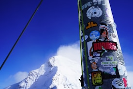 A snow-covered mountain peak under a clear blue sky with a foreground featuring a pole covered in various colorful stickers, including logos and brand names. A cable runs diagonally across the scene, suggesting a ski lift or similar structure.