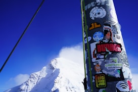 A snow-covered mountain peak under a clear blue sky with a foreground featuring a pole covered in various colorful stickers, including logos and brand names. A cable runs diagonally across the scene, suggesting a ski lift or similar structure.