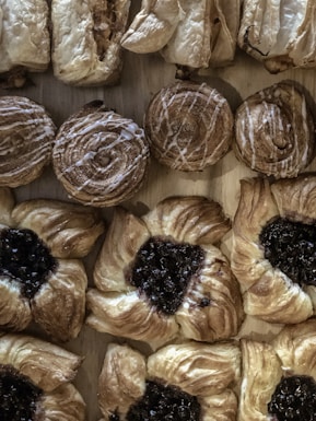 An assortment of pastries is arranged neatly on a wooden surface. The variety includes round pastries with icing drizzles, flaky rolls with visible layers, and pastries with a dark fruit filling in the center. The textures of the pastries range from smooth icing to crumbly, crisp layers.
