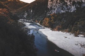 A serene river meanders through a lush forested landscape. Cliffs rise on one side, covered in dense, dark green foliage, while the riverbank displays a sandy, pebbled beach. A person in a kayak can be seen paddling along the calm water, adding a sense of adventure and tranquility to the scene.