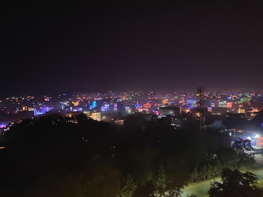 A nighttime cityscape with buildings illuminated in various vibrant colors, creating a lively and festive atmosphere across the horizon. A silhouette of dark trees or foliage appears in the foreground, adding depth to the image. The sky is dark, highlighting the colorful lights of the buildings.