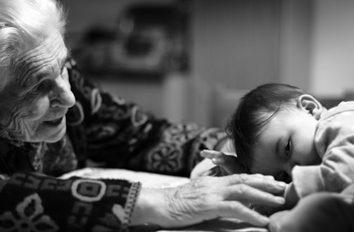 An elderly person with white hair leans towards a baby, smiling warmly. Their hands gently touch, conveying a sense of connection and tenderness. The black-and-white setting enhances the emotional depth of the moment.