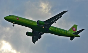 A green commercial airplane with the logo S7 on its body and tail is flying against a cloudy sky. The aircraft is in mid-flight with its landing gear extended, indicating either a landing or takeoff phase.