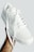 A white sneaker is held in a hand, featuring perforated details on the side and a clean, minimalist design. The shoelaces are neatly tied and dangle freely. The background is a soft, muted tone, creating a simple and modern aesthetic.