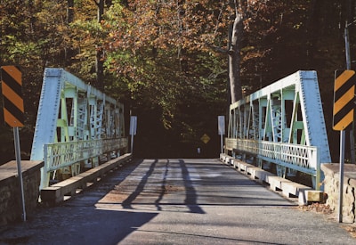 A narrow metal bridge with steel beams and railings crosses a paved road. The structure is flanked by stone parapets and surrounded by a dense forest of trees with autumn foliage. Yellow and black striped warning signs are positioned at the entrance, and shadows from the bridge fall across the pavement.