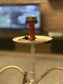 A hookah with a red bowl and burning coals sits on a white tray. The background features a blurred television screen and a brick wall, creating a cozy and relaxed ambiance.