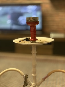A hookah with a red bowl and burning coals sits on a white tray. The background features a blurred television screen and a brick wall, creating a cozy and relaxed ambiance.