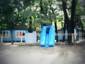A collection of small, blue, wooden cabins situated among trees, with a blue fence in front. There are clothes hanging on the fence, including a red and a black article. Bright blue kayaks are stacked vertically against the fence.